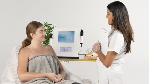 A beautician in a white uniform talks to a client wrapped in a gray towel at a skincare clinic, with a facial treatment device in the background.
