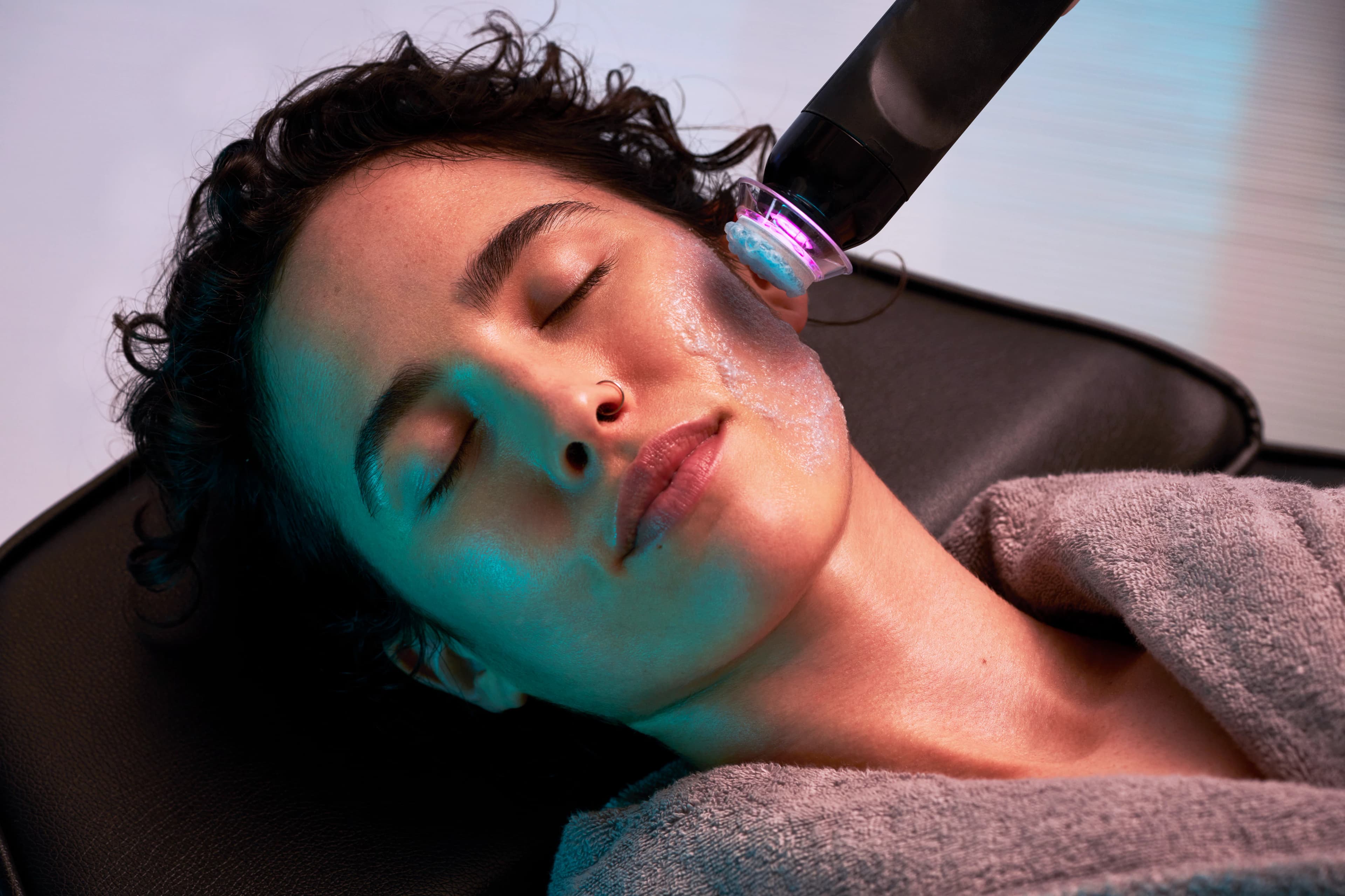 A woman relaxes with her eyes closed on a treatment bed while a provider uses the Glo2Facial handpiece on her cheek.