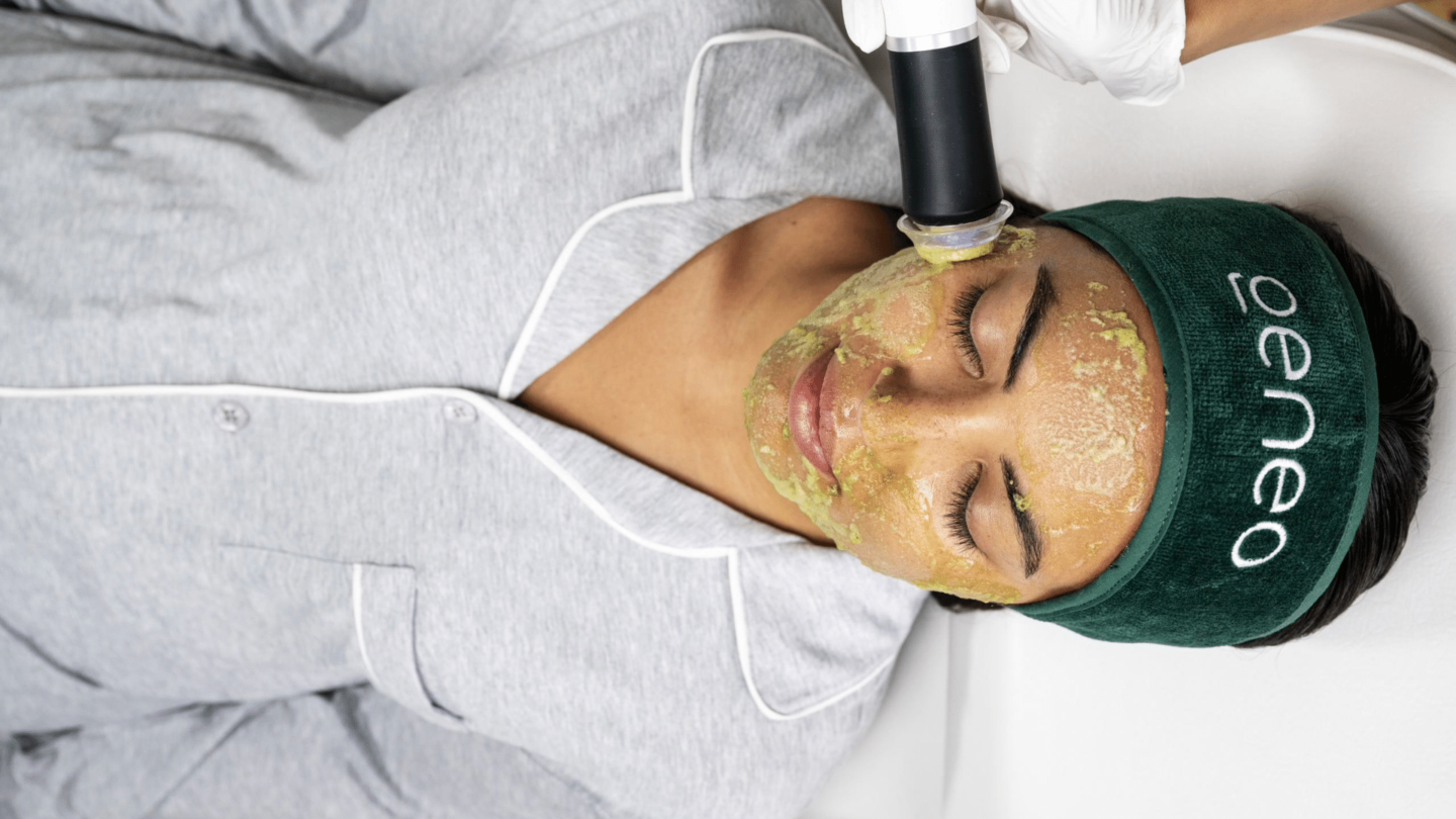 Woman in grey shirt enjoying a facial massage with a Geneo facial treatment, wearing a green Geneo headband or hair towel