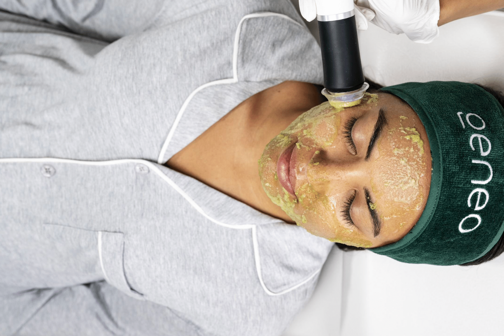 Woman in grey shirt enjoying a facial massage with a Geneo facial treatment, wearing a green Geneo headband or hair towel