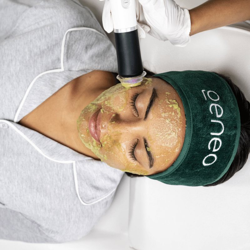 Woman receiving a Geneo Detox facial treatment, wearing a dark green Geneo-branded headband