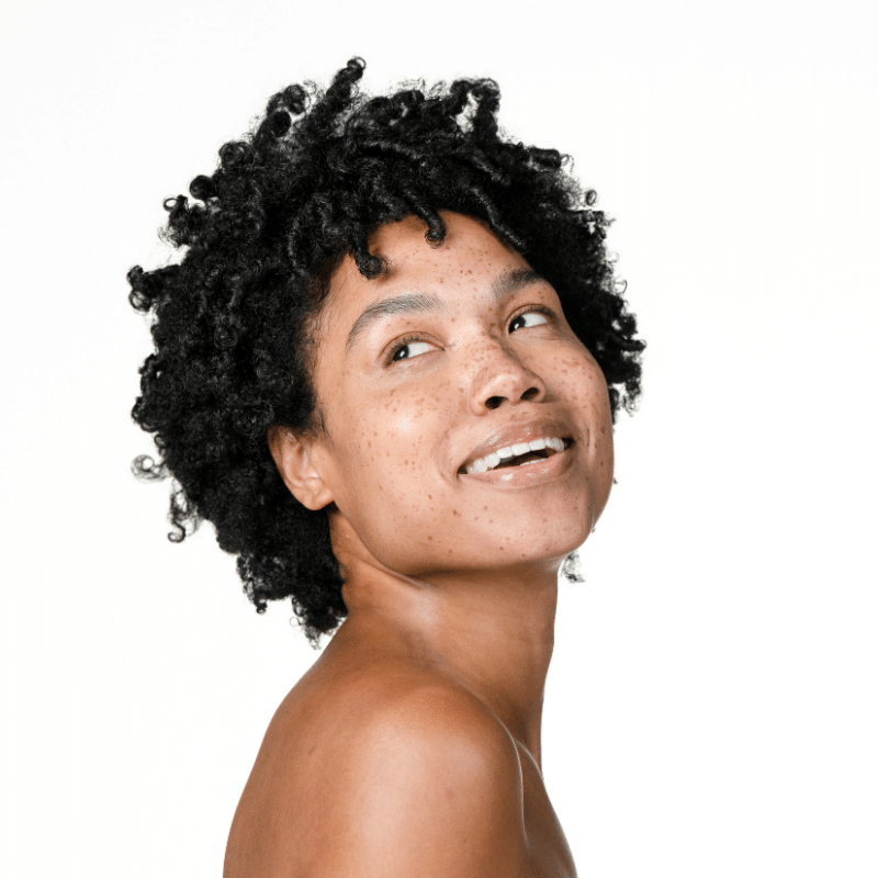 Woman with dark coily hair and freckled, glowing skin smiling and looking over her shoulder